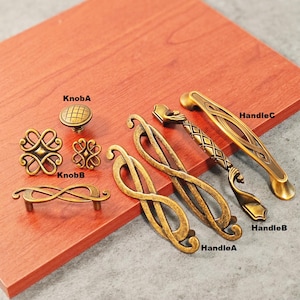 May include: A collection of antique brass cabinet hardware, including knobs and handles. The knobs feature a grid pattern and a decorative floral design. The handles have intricate scroll and geometric patterns. The hardware is displayed on a reddish-brown surface.