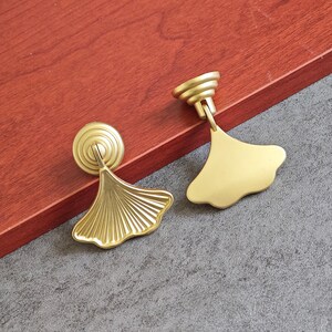 May include: Two brass cabinet pulls with a ginkgo leaf design. The pulls have a circular base and a fan-shaped leaf, with a gold finish. The pulls are set against a red-brown wooden surface and a gray surface.