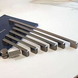 Może przedstawiać: Zestaw nowoczesnych, ciemnoszarych uchwytów i gałek do szafek. Uchwyty są prostokątne z zakrzywionym uchwytem, a gałki są okrągłe i prostokątne. Okucia są ułożone na białej powierzchni z ciemnoniebieską teczką w tle.