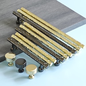 May include: A collection of cabinet hardware, including gold and black hammered bar pulls and matching round knobs. The pulls have a textured surface and are arranged in a diagonal pattern. The knobs are displayed in the foreground, showcasing their design.