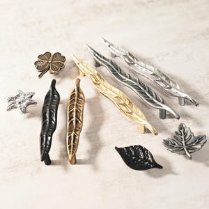 May include: A selection of decorative cabinet hardware, including leaf-shaped and clover-shaped knobs and pulls. The hardware is in various metallic finishes, such as gold, silver, and black. The hardware is designed for furniture and cabinetry.