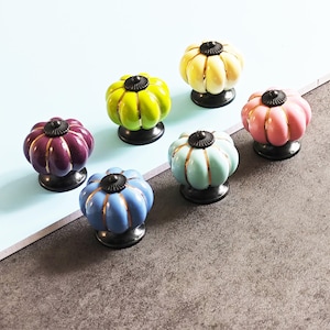 May include: A collection of six colorful ceramic cabinet knobs. Each knob is shaped like a pumpkin with a black base and a decorative gold accent. The knobs are in shades of purple, green, yellow, pink, blue, and teal.