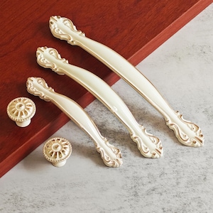 Könnte beinhalten: Cremefarbenes Schrankbeschlagsset mit goldfarbenem Besatz. Das Set enthält zwei dekorative Knöpfe und drei Griffleisten in verschiedenen Größen. Die Beschläge haben ein verziertes, Vintage-Design, passend für Küchen- oder Badezimmerschränke.