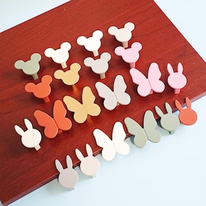 Könnte beinhalten: Eine Sammlung farbenfroher Schrankknöpfe in verschiedenen Formen, darunter Mickey Mouse-Ohren, Schmetterlinge und Hasen. Die Knöpfe sind in Weiß-, Rosa-, Orange-, Grün- und Beigetönen auf einer dunkelroten Holzoberfläche angeordnet.