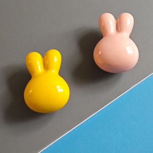 May include: Two glossy, bunny-shaped decorative objects. One is bright yellow, the other is soft pink. The objects have a rounded body with two upright, rounded ears. They are set against a grey and blue background.