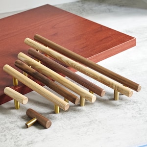 May include: A selection of wooden cabinet pulls in shades of brown and tan. Each pull has a cylindrical wooden handle and a brass-coloured base. The pulls are arranged on a reddish-brown surface.