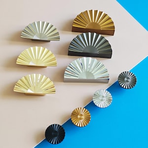 Może przedstawiać: Kolekcja dekoracyjnych okuć do szafek w różnych metalicznych wykończeniach, w tym złotym, srebrnym i czarnym. Projekty charakteryzują się kształtem wachlarza i okrągłymi gałkami, ułożonymi na różowym i niebieskim tle.