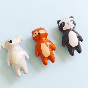 Könnte beinhalten: Drei Keramik-Tierfiguren: ein Koala, ein Fuchs und ein Dachs. Der Koala ist hellgrau mit cremefarbenem Bauch, der Fuchs ist orange mit cremefarbenem Bauch und der Dachs ist schwarz und weiß. Alle haben glänzende Oberflächen und sind vor hellblauem Hintergrund platziert.