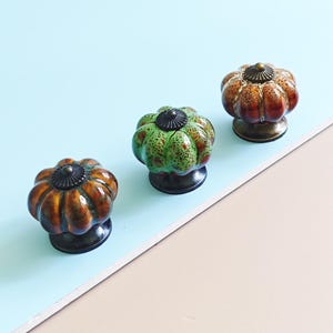 May include: Three decorative cabinet knobs in a pumpkin shape. The knobs are in shades of orange, brown, and green with a dark bronze base and top. The knobs are arranged on a light blue and beige surface.