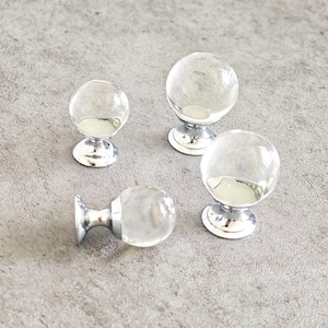 Peut inclure: Quatre boutons de meuble en verre transparent, de forme sphérique, avec des bases argentées. Les boutons sont disposés sur une surface grise, mettant en valeur leurs qualités réfléchissantes et leur élégance simple. Idéal pour les meubles.