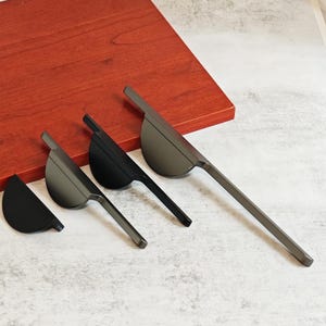 May include: Four modern cabinet pulls in varying shades of gray and black. Each pull features a curved, fan-shaped handle attached to a slender, rectangular bar. The pulls are arranged diagonally against a light gray surface and a red-brown wooden background.