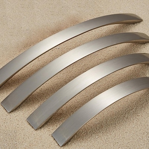 Op de afbeelding: Vier geborsteld nikkel kastgrepen in verschillende maten zijn diagonaal gerangschikt. Elke greep heeft een gebogen, ergonomisch ontwerp met een rechthoekige basis. De grepen zijn gemaakt van metaal en hebben een matte afwerking.