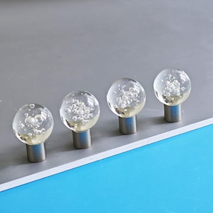 May include: Four clear glass cabinet knobs with a bubble design. Each knob has a silver base and a round, transparent glass top filled with small, air-like bubbles. The knobs are arranged in a row on a grey surface.
