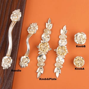 Op de afbeelding: Een verzameling zilverkleurige kastbeslagen, waaronder een handgreep, knoppen en platen, allemaal met een rozenontwerp. De hardware wordt gepresenteerd tegen een oranje achtergrond en een houten oppervlak.