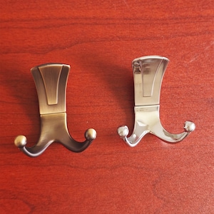 May include: Two metal coat hooks. One has a bronze finish, the other is silver. Each hook features a decorative top and two curved arms with ball-shaped ends. They are displayed on a reddish-brown wooden surface.