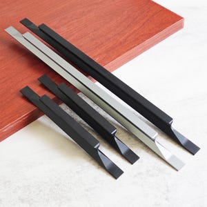 Può includere: Una collezione di maniglie per mobili moderne in nero e argento. Le maniglie hanno un design a barra rettangolare con un bordo smussato. L'hardware è esposto su una superficie di legno marrone rossiccio.