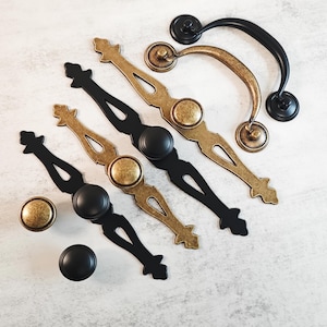 Könnte beinhalten: Eine Sammlung von Schrankbeschlägen, darunter Knöpfe und Griffe in Schwarz und Antik-Messing. Die Griffe haben dekorative Designs und sind in verschiedenen Größen erhältlich. Die Knöpfe sind rund und schlicht. Die Hardware ist auf einer hellen Oberfläche angeordnet.