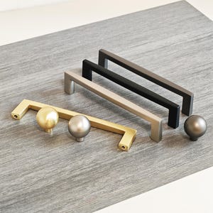 Könnte beinhalten: Eine Auswahl an Schrankbeschlägen, darunter goldene, silberne, schwarze und graue Griffe und Knöpfe. Die Griffe sind rechteckig und die Knöpfe kugelförmig. Die Beschläge werden auf einer grauen Oberfläche präsentiert.