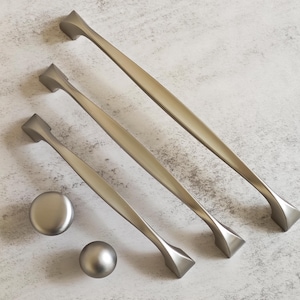 Könnte beinhalten: Eine Auswahl an Schrankbeschlägen aus gebürstetem Nickel. Das Bild zeigt drei unterschiedlich große Griffe und zwei Knöpfe. Die Griffe haben ein modernes Design mit einem gebogenen Griff und geometrischen Enddetails. Die Knöpfe sind rund und kugelförmig.