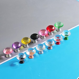 Pode incluir: Uma coleção de puxadores de armário de cristal coloridos. Os puxadores apresentam tampas facetadas em forma de gema em várias cores, incluindo rosa, roxo, amarelo, preto, vermelho, verde e transparente. Cada puxador tem uma base prateada.