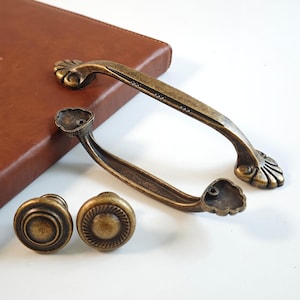 Op de afbeelding: Antiek bronzen kastbeslagset, bestaande uit twee handgrepen en twee knoppen. De handgrepen hebben een gebogen ontwerp met decoratieve bloemaccenten. De knoppen hebben een cirkelvormig ontwerp met concentrische cirkels.