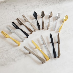 May include: A collection of decorative cabinet hardware featuring spoon, fork, and knife designs. The hardware is in various colors, including gold, black, silver, and bronze. The handles are designed to resemble silverware.