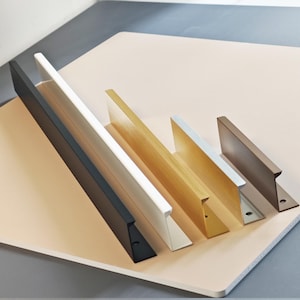 Può includere: Una collezione di maniglie per mobili in vari colori, tra cui nero, bianco, oro e marrone. Ogni maniglia ha un design elegante e moderno con una parte anteriore piatta e un'impugnatura ad angolo retto. Sono disposte su una superficie rosa chiaro.