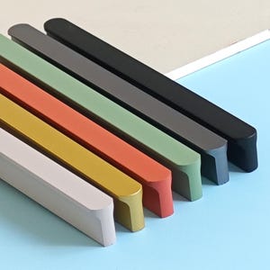 May include: A collection of six rectangular cabinet pulls in various colors, including off-white, yellow, orange, green, gray, and black. The pulls are arranged diagonally against a light blue surface.