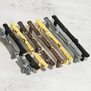 Pode incluir: Uma coleção de puxadores decorativos para armários em vários acabamentos metálicos, incluindo prata, preto, ouro e bronze. Os puxadores têm um design texturizado em forma de ramo, adicionando uma estética natural e rústica. Estas peças de hardware são adequadas para gavetas e armários.