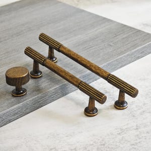 May include: A collection of antique brass cabinet handles and a knob. The set includes two bar pulls with a ribbed central design and a round knob. The hardware is displayed on a grey surface. Ideal for furniture.