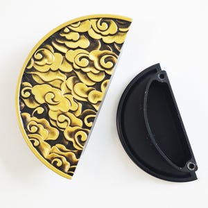 May include: Two half-circle cabinet pulls. One is gold with a cloud design, the other is black. The gold pull has a detailed relief pattern. The black pull is a simple, solid colour. Both are likely for furniture.