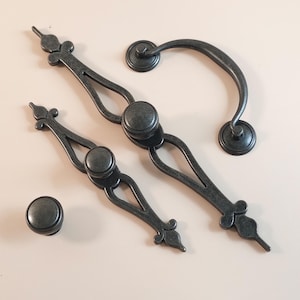 Op de afbeelding: Een verzameling antieke meubelbeslag in een donkere, verweerde metalen afwerking. De set bevat twee langwerpige grepen, een gebogen handgreep en een ronde knop. Het beslag heeft decoratieve details en een vintage esthetiek.