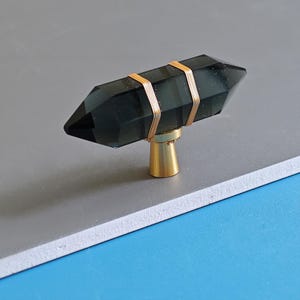 May include: A dark grey crystal cabinet knob with gold wire detailing. The knob has a gold-coloured base. The crystal is a dark, translucent colour. The knob is designed for use on cabinets and drawers.
