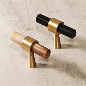 Pode incluir: Dois puxadores de armário com design cilíndrico. Um tem o centro preto, o outro castanho claro. Ambos os puxadores têm um anel e uma base dourados, criando um visual moderno e elegante. Os puxadores são projetados para fácil instalação.