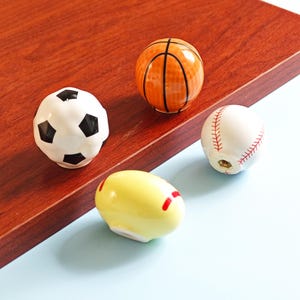 Op de afbeelding: Een verzameling keramische kastknoppen in de vorm van sportballen. Inclusief een voetbal, basketbal, honkbal en een gele eivormige knop met rode strepen. De knoppen liggen op een houten oppervlak.