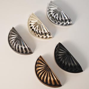 May include: A selection of half-moon cabinet handles in diverse metallic finishes: silver, gold, bronze, and black. Each handle has a fan-shaped design with detailed ridges. These offer a decorative and functional hardware option for furniture.