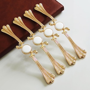 May include: Six gold cabinet pulls with a decorative design. Each pull has a white circular centre and a stylized, fan-shaped end. The pulls are arranged on a white surface with a brown leather-like background.