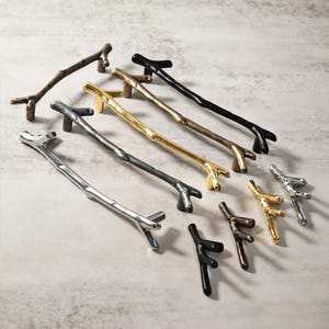 May include: A selection of branch-shaped cabinet pulls in diverse metallic finishes, including silver, gold, bronze, and black. The hardware features a natural twig design, suitable for furniture or cabinetry. The pulls vary in size and shape.