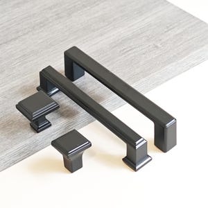 Może przedstawiać: Zestaw czterech czarnych elementów okuć do szafek. Zestaw zawiera dwa kwadratowe gałki i dwa prostokątne uchwyty. Okucia wykonane są z matowego czarnego materiału i są prezentowane na jasnoszarej powierzchni.