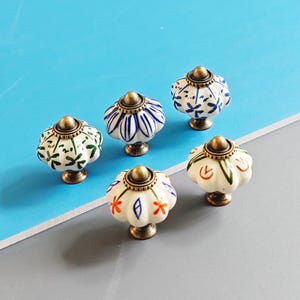 May include: A collection of five decorative cabinet knobs. Each knob features a white ceramic floral design with brass hardware. The knobs have different floral patterns in blue, green, orange, and white.