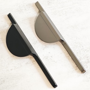 May include: Two contemporary cabinet pulls, one black and one grey, are shown against a light backdrop. Each pull has a semi-circular handle connected to a slim, rectangular bar. The hardware is designed for easy installation and a sleek aesthetic.