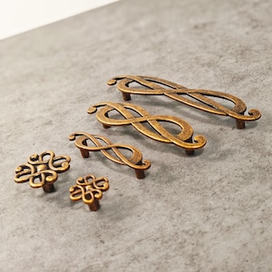 Può includere: Una collezione di ferramenta per mobili in bronzo antico, tra cui maniglie e pomelli. Le maniglie presentano un design a simbolo dell'infinito, mentre i pomelli hanno un motivo ornato e a vortice. L'hardware è esposto su una superficie grigia.