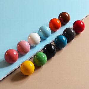 Könnte beinhalten: Eine Sammlung von zwölf glänzenden, kugelförmigen Knöpfen in verschiedenen Farben, darunter Rot, Rosa, Gelb, Grün, Blau, Schwarz und Braun. Die Knöpfe sind diagonal vor einem zweifarbigen blau-beige Hintergrund angeordnet.