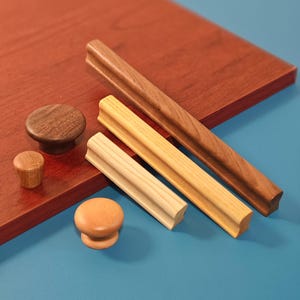 May include: A selection of wooden cabinet hardware, including knobs and pulls. The knobs are round, with one dark brown and two light brown. The pulls are rectangular, with a dark brown and two light brown. The hardware is displayed on a red-brown surface.