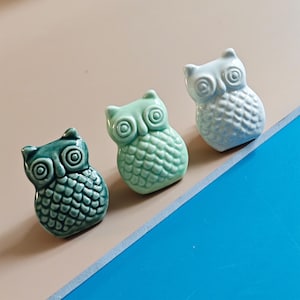 Könnte beinhalten: Drei Keramik-Eulenfiguren in den Farbtönen Türkis, Grün und Hellblau. Jede Eule hat große, runde Augen und einen strukturierten Körper. Die Figuren sind diagonal vor einem neutralen Hintergrund angeordnet.