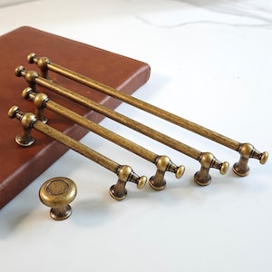 Op de afbeelding: Set antieke messing kastbeslag, bestaande uit vier stanggrepen en een ronde knop. De grepen hebben een klassiek ontwerp met decoratieve uiteinden. De hardware wordt gepresenteerd op een wit oppervlak met een bruine achtergrond.