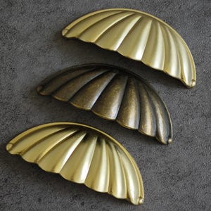 Peut inclure: Trois poignées de meubles décoratives en forme d'éventail ou de coquillage. Deux sont de couleur or vif et une est d'un bronze plus foncé. Les poignées ont un design strié et sont en métal. Le fond est gris foncé.