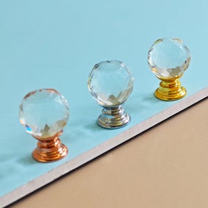 May include: Three crystal ball cabinet knobs with a faceted design. The knobs have metallic bases in gold, silver, and copper colours. The knobs are displayed on a light blue and tan surface.