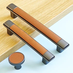 May include: A set of cabinet handles and a knob. The handles are rectangular with a dark grey metal frame and a textured brown leather-effect centre. The knob is round with a matching design. Suitable for furniture.
