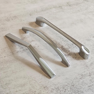 Pode incluir: Três puxadores de armário prateados com um design moderno. Os puxadores têm uma forma curva ou angular e são feitos de um material metálico brilhante. Estão dispostos sobre uma superfície cinza claro, mostrando sua estética elegante e contemporânea.